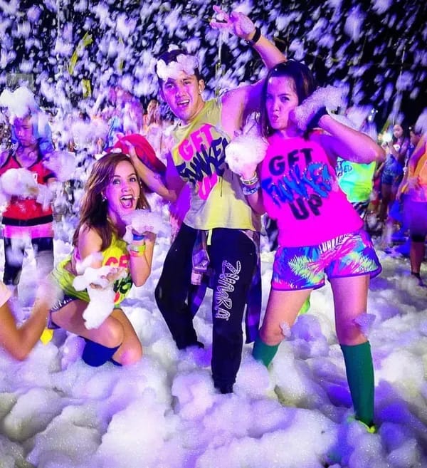 Adults in neon shirts enjoying a foam party at night under colorful lights